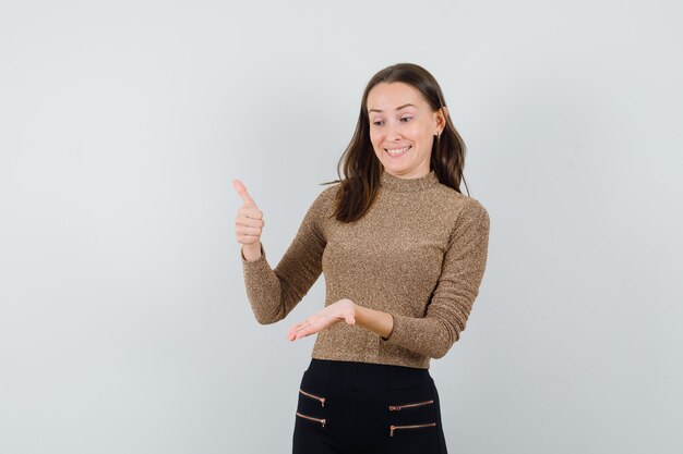 Jonge vrouw in goud vergulde trui en zwarte broek duim opdagen en doen alsof ze naar de telefoon kijken en gelukkig, vooraanzicht kijken.