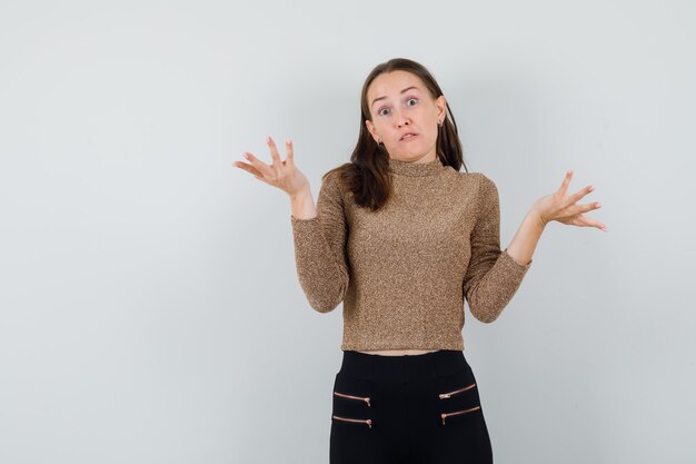 Jonge vrouw in goud vergulde trui en zwarte broek schouders ophalen en op zoek peinzend, vooraanzicht.