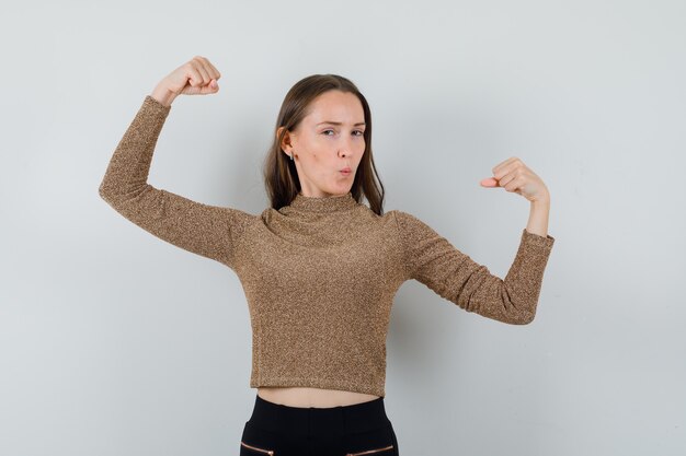 Jonge vrouw in gouden blouse die haar armen opheft terwijl ze haar kracht toont en er indrukwekkend uitziet, vooraanzicht.