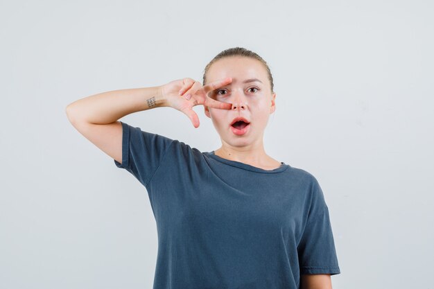 Jonge vrouw in grijs t-shirt dat v-teken dichtbij oog toont en verbaasd kijkt