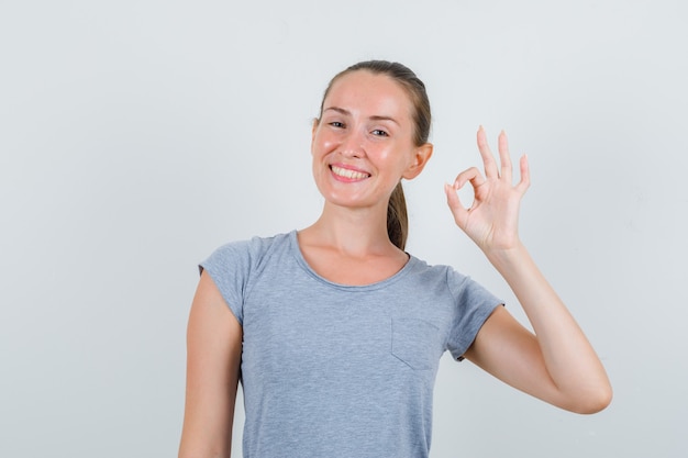 Jonge vrouw in grijs t-shirt die ok gebaar toont en gelukkig, vooraanzicht kijkt.