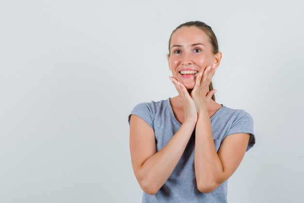 Jonge vrouw in grijs t-shirt kin aan te raken en op zoek vrolijk, vooraanzicht.