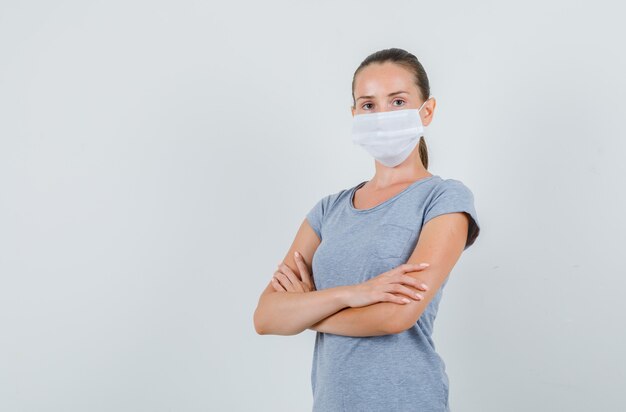 Jonge vrouw in grijs t-shirt, masker staande met gekruiste armen en op zoek zelfverzekerd, vooraanzicht.