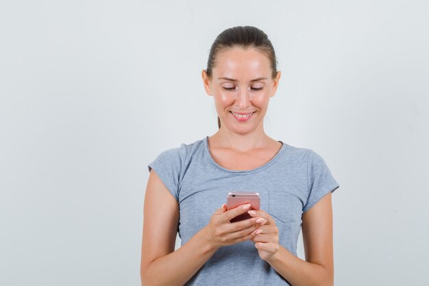 Jonge vrouw in grijs t-shirt met behulp van mobiele telefoon en op zoek vrolijk, vooraanzicht.