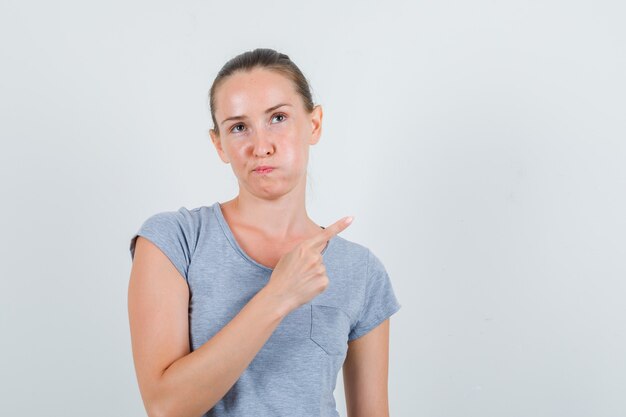 Jonge vrouw in grijs t-shirt wijst naar kant en kijkt ontevreden, vooraanzicht.