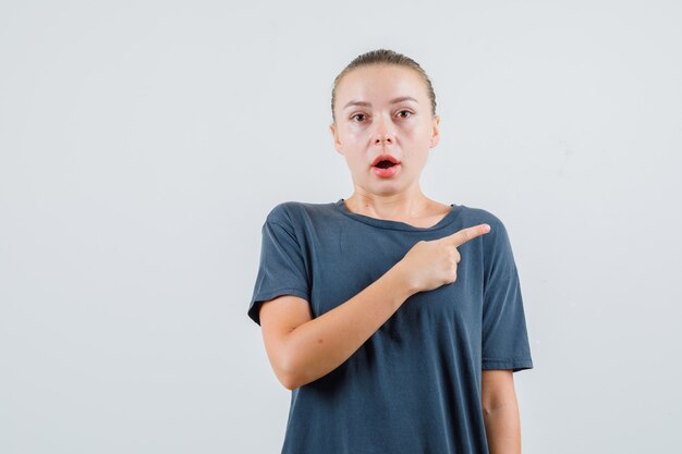 Jonge vrouw in grijs t-shirt wijst naar kant en kijkt verbaasd