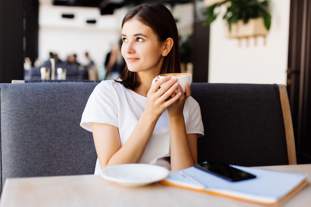 Jonge vrouw in grijze jurk zittend aan tafel in café en schrijven in notitieblok. Studenten leren online.