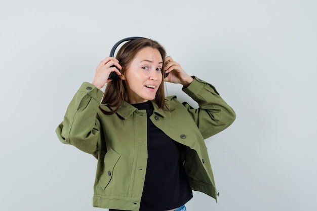 Jonge vrouw in groen jasje die oortelefoons draagt en tevreden, vooraanzicht kijkt.