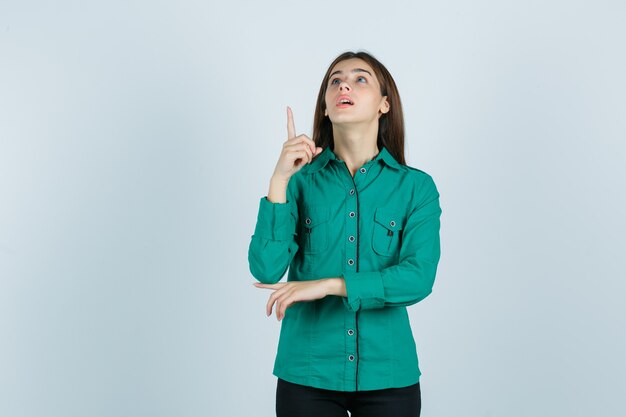 Jonge vrouw in groen shirt die naar boven wijst en vraagt zich af, vooraanzicht.