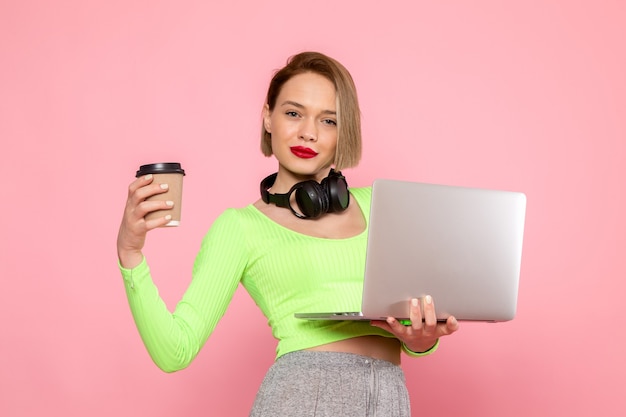 Jonge vrouw in groen shirt en grijze broek poseren met laptop en zwarte koptelefoon