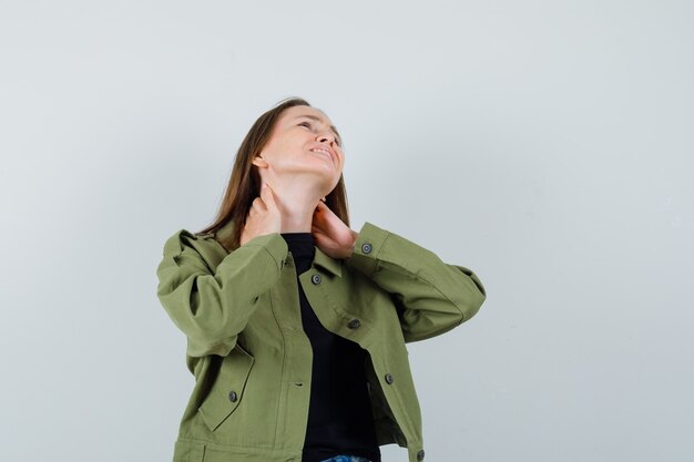 Jonge vrouw in groene jas die aan nekpijn lijdt en ongemakkelijk, vooraanzicht kijkt.