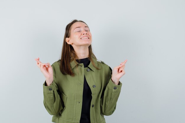 Jonge vrouw in groene jas die zich met gekruiste vingers bevindt en wenselijk, vooraanzicht kijkt.