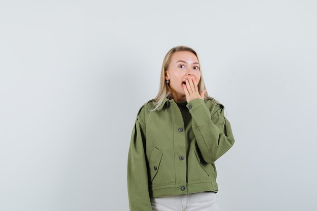 Jonge vrouw in groene jas, jeans met hand op de mond terwijl lachen en kijken verbaasd, vooraanzicht.