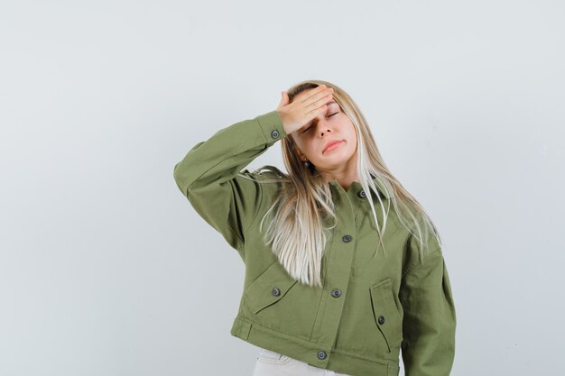 Jonge vrouw in groene jas met hand op haar voorhoofd en op zoek moe, vooraanzicht.