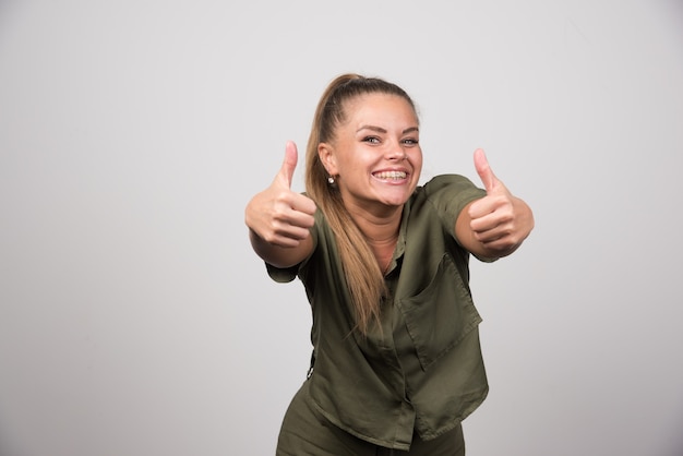 Jonge vrouw in groene trui duimen opgevend op grijze muur.