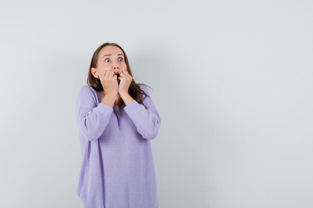Jonge vrouw in lila blouse met vuisten op haar mond en bang op zoek