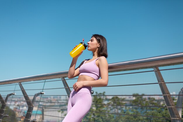 Jonge vrouw in montage sport slijtage op brug op warme zonnige ochtend met fles water shaker dorstig na training moe drinken