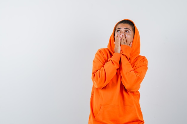 Jonge vrouw in oranje hoodie die mond bedekt met handen en verbaasd kijkt