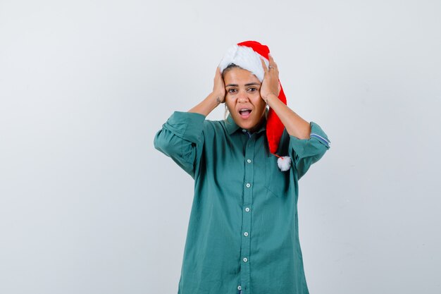 Jonge vrouw in overhemd, kerstmuts die de handen op het hoofd houdt en er geschokt uitziet, vooraanzicht.