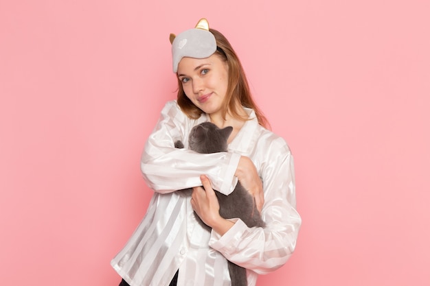 jonge vrouw in pyjama's en slaapmasker grijs kitten op roze te houden