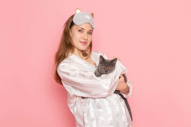 Jonge vrouw in pyjama's en slaapmasker poseren met glimlach en grijs kitten op roze