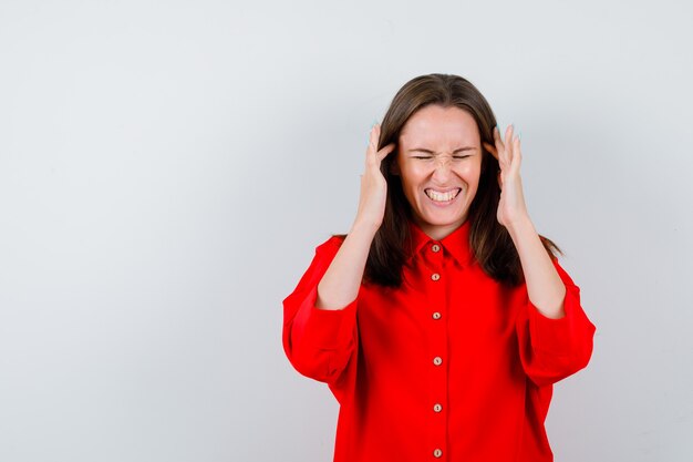 Jonge vrouw in rode blouse met vingers op tempels en op zoek pijnlijk, vooraanzicht.