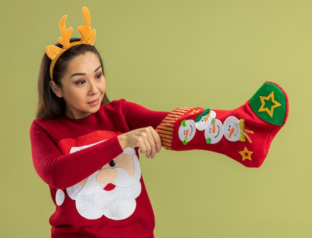 Jonge vrouw in rode kersttrui met grappige rand met hertenhoorns die kerstsok vasthoudt en er geïntrigeerd uitziet over groene muur