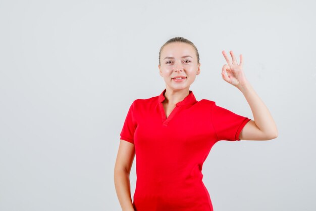 Jonge vrouw in rode t-shirt die ok gebaar toont en gelukkig kijkt
