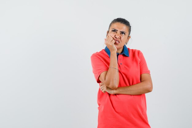 Jonge vrouw in rode t-shirt leunende hand op de wang, staande in denken pose en peinzend op zoek, vooraanzicht.