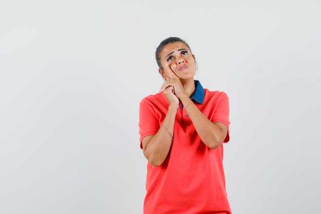 Jonge vrouw in rode t-shirt leunende wang aan kant en peinzend, vooraanzicht kijkt.