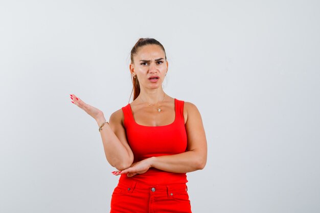 Jonge vrouw in rood mouwloos onderhemd, broek die handpalm uitspreidt en wrokkig, vooraanzicht kijkt.