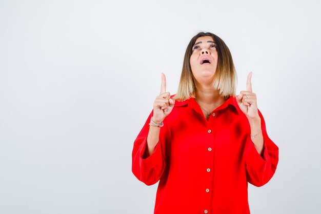 Jonge vrouw in rood oversized shirt die omhoog wijst en verbaasd kijkt, vooraanzicht.