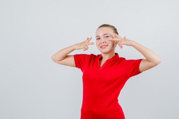 Jonge vrouw in rood t-shirt dat v-teken dichtbij ogen toont en gelukkig kijkt