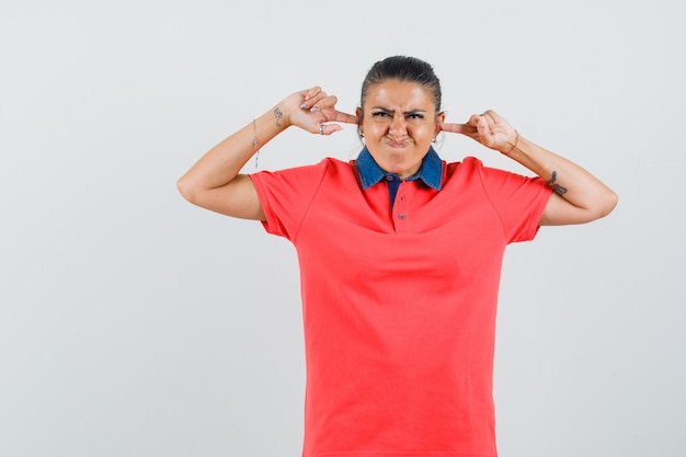 Jonge vrouw in rood t-shirt oren met wijsvingers inpluggen en geërgerd, vooraanzicht kijken.