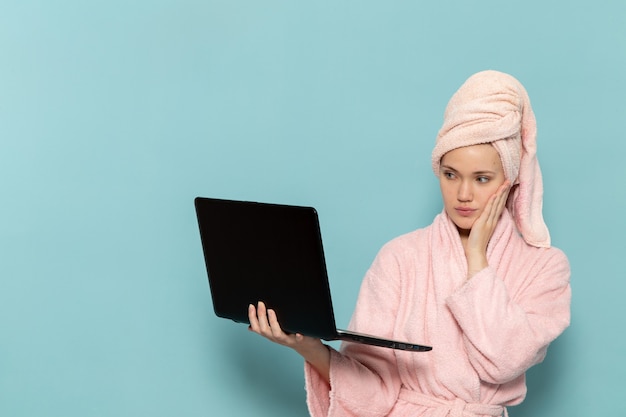 jonge vrouw in roze badjas na het douchen met donkere laptop op blauw bureau