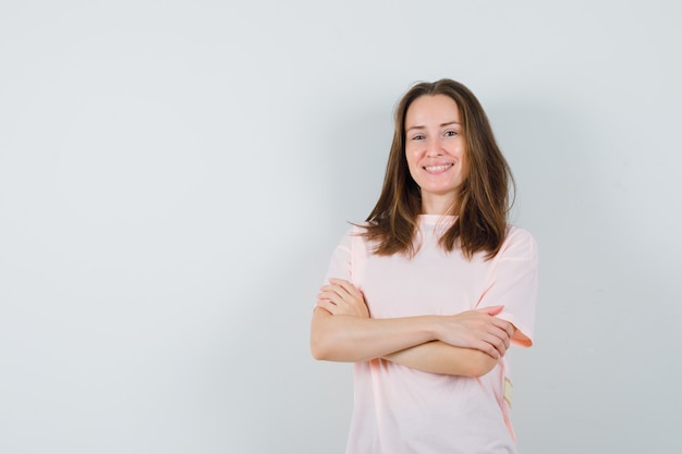 Jonge vrouw in roze t-shirt staande met gekruiste armen en op zoek zelfverzekerd, vooraanzicht.