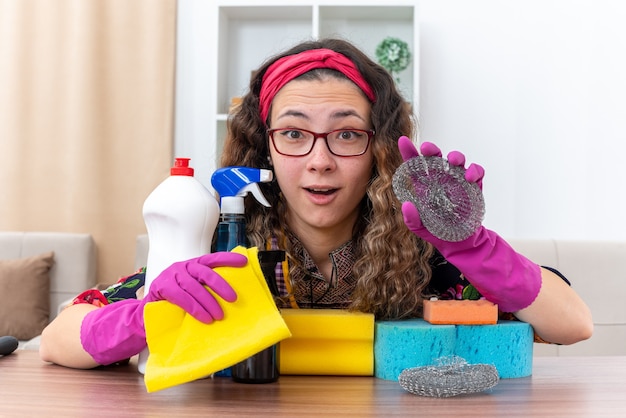 Jonge vrouw in rubberen handschoenen camera kijken verbaasd en gelukkig zitten aan de tafel met schoonmaakbenodigdheden en gereedschappen in lichte woonkamer