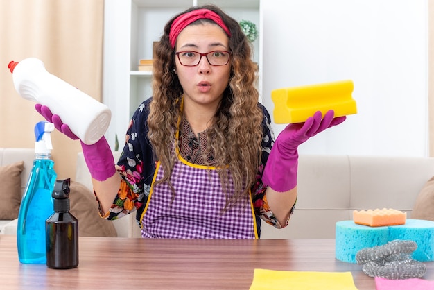 Jonge vrouw in rubberen handschoenen met spons en fles met schoonmaakspullen en kijkt verrast aan tafel met schoonmaakspullen en gereedschap in lichte woonkamer