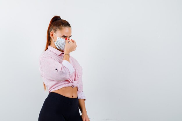 Jonge vrouw in shirt, broek, medisch masker neusbrug masseren en op zoek uitgeput, vooraanzicht.