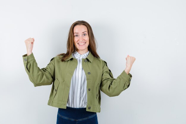 Jonge vrouw in shirt, jas die winnaargebaar toont en er gelukkig uitziet, vooraanzicht.