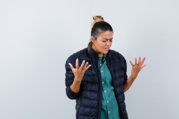 Jonge vrouw in shirt, pufferjack die handen opsteekt, naar beneden kijkt en weemoedig kijkt, vooraanzicht.