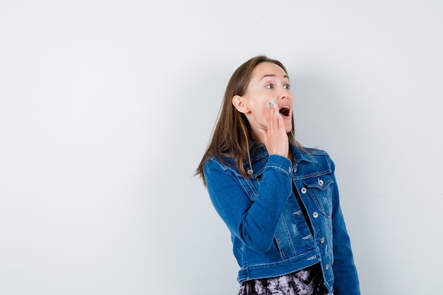 Jonge vrouw in spijkerjasje, jurk schreeuwt door hand in de buurt van mond te houden en opgewonden te kijken, vooraanzicht.