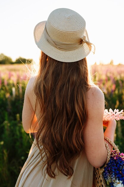 Jonge vrouw in strohoed en kleding met boeket van lupinebloemen