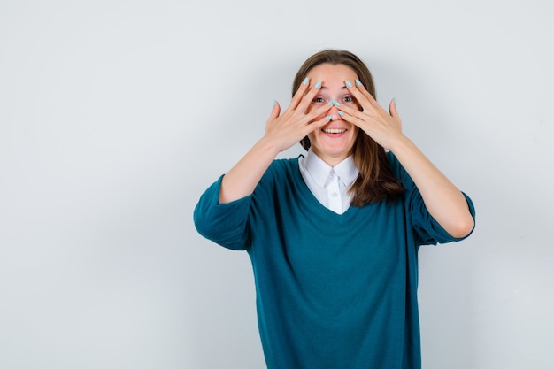 Jonge vrouw in sweater over wit overhemd die door vingers kijkt en gelukkig, vooraanzicht kijkt.