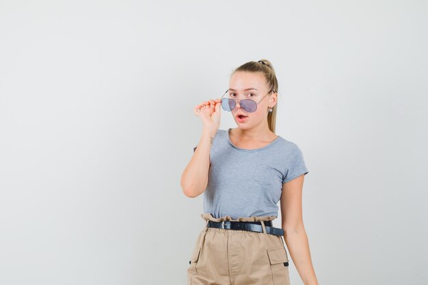 Jonge vrouw in t-shirt, broek kijkt uit over een bril en kijkt verbaasd, vooraanzicht.