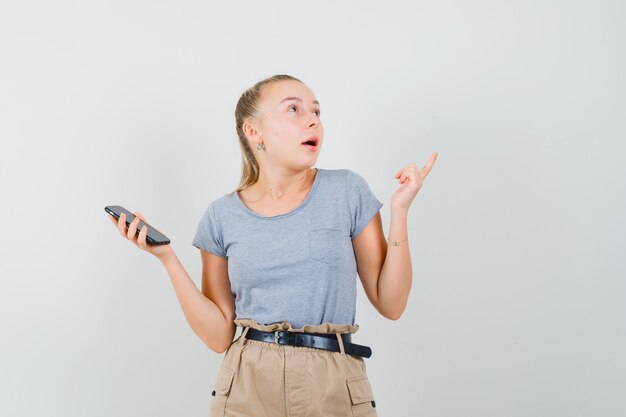 Jonge vrouw in t-shirt, broek met mobiele telefoon, omhoog en op zoek verbaasd, vooraanzicht.