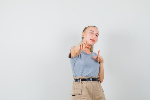 Jonge vrouw in t-shirt, broek wijst naar voren en kijkt zelfverzekerd, vooraanzicht.