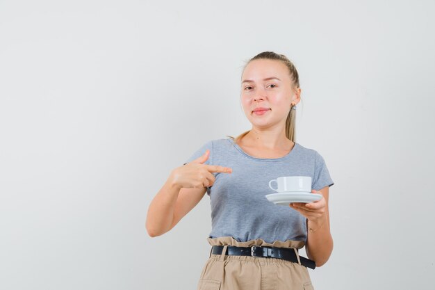 Jonge vrouw in t-shirt, broek wijzend op kopje drank en kijkt blij, vooraanzicht.