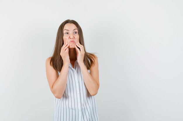 Jonge vrouw in t-shirt die de huid van het gezicht op agressieve wijze naar beneden trekt en zeer gespannen, vooraanzicht kijkt.