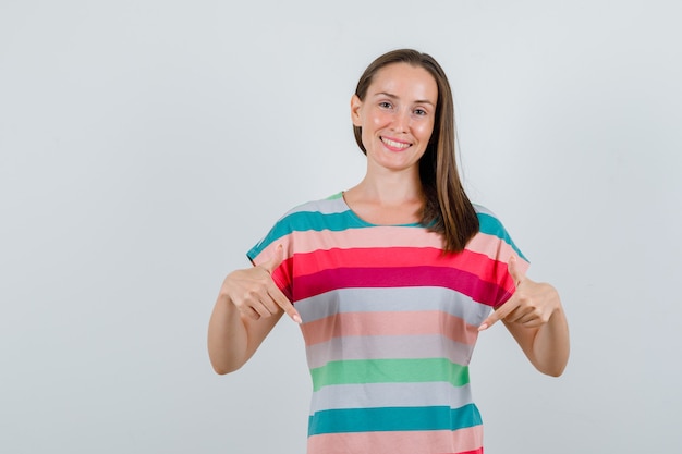 Jonge vrouw in t-shirt die naar beneden wijst en vrolijk, vooraanzicht kijkt.